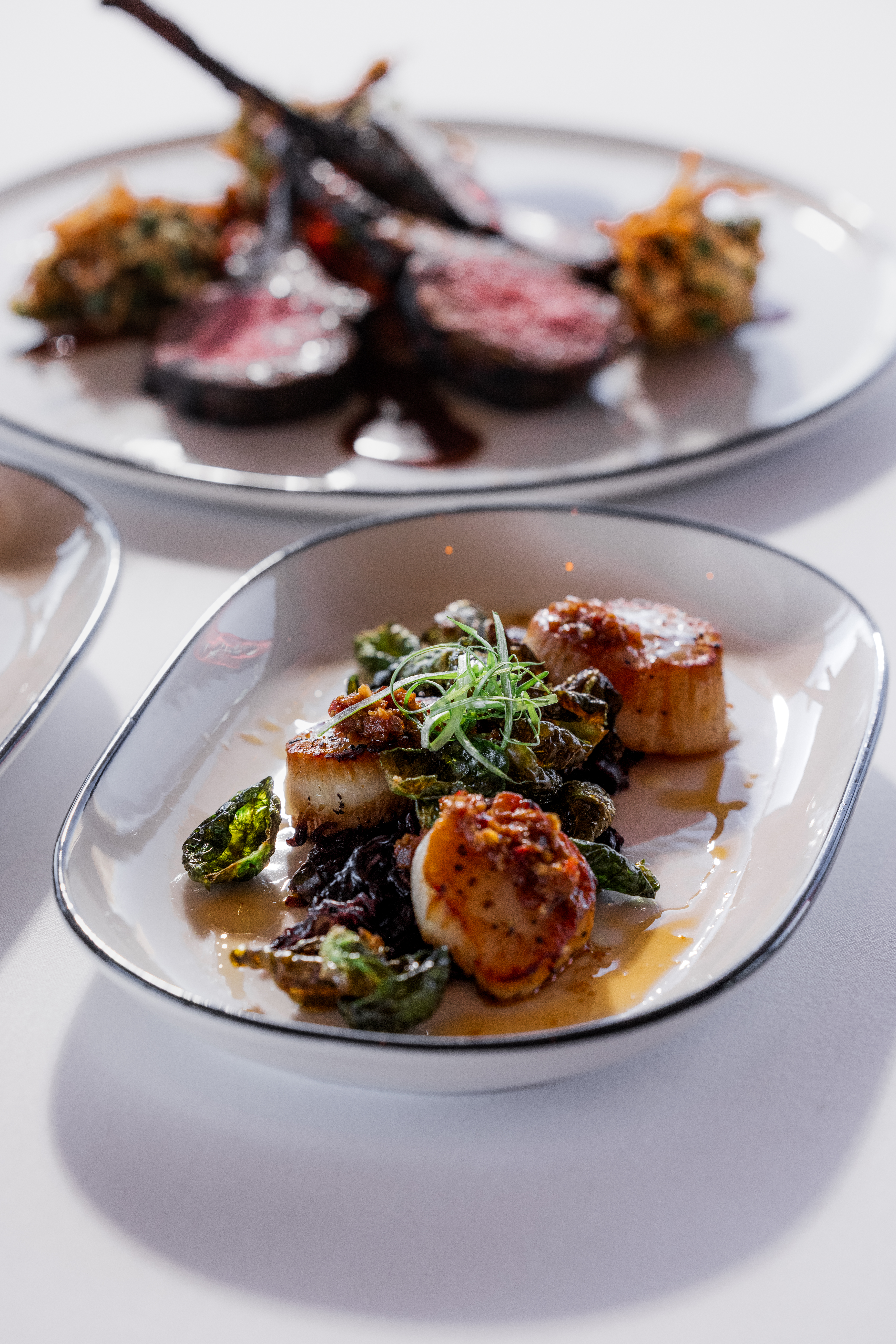 pan fried scallops with XO sauce on fried brussels sprouts in front of a plate with lamb chops with a side of pakora